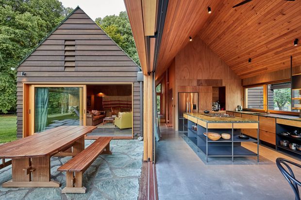 A warm, contemporary indoor-outdoor living and kitchen space in a forested setting. The image shows a timber-clad gabled pavilion with large sliding glass openings, a stone-paved outdoor dining area, and an open-plan kitchen with a timber-lined vaulted ceiling, timber cabinetry, a central island, and polished concrete flooring. A living room is visible through the glazing.