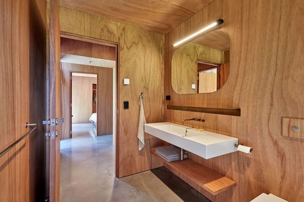 Warm contemporary bathroom clad extensively in natural timber veneer, featuring a wall-mounted white basin, black tapware, rounded rectangular mirror with integrated lighting, open timber shelving, and a timber bench beneath the vanity. A timber-framed doorway opens to a bedroom or adjoining room.
