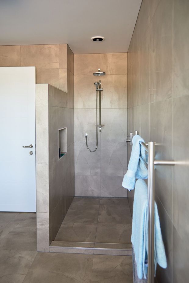 A modern walk-in shower bathroom with large-format beige and grey stone-look tiles covering the walls and floor. The shower features a rain showerhead, handheld shower, recessed wall niche, frameless glass screen, and chrome fixtures. A white door and chrome towel rail with light blue towels are visible.