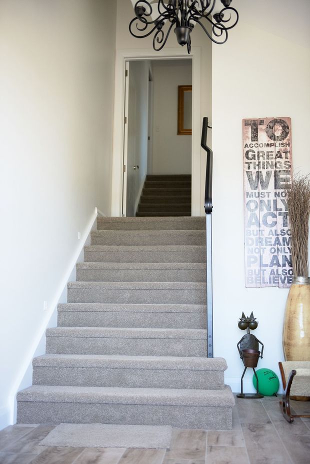 A bright residential stairway viewed from the lower level, with broad grey carpeted steps, a dark metal handrail, white walls, and a decorative black chandelier overhead. The stairwell leads to an upper hallway, with a framed artwork, tall floor vase, and small decorative objects at the side. The lower floor is finished in light timber-look or stone-look planks.