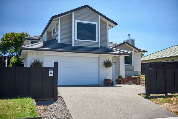 Exterior view of a contemporary two-storey suburban home with a gabled upper level, light horizontal weatherboard cladding, pale masonry around the garage and entry, dark tiled roofing, and a wide double garage. A paved driveway leads through a dark privacy fence and gate.