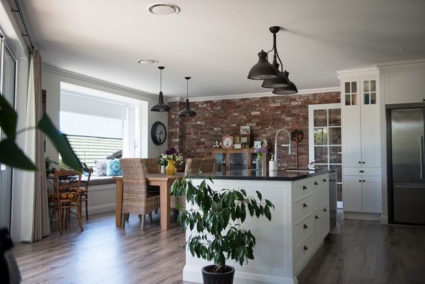 Bright open-plan kitchen and dining area with a large white island, shaker-style cabinetry, dark pendant lights, timber-look flooring, and a red exposed-brick feature wall. A dining table sits beside large windows, with a stainless-steel refrigerator and glass-front storage cabinetry visible at right. Indoor plants and natural light create a warm, inviting atmosphere.