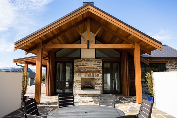 Exterior view of a rustic mountain-style home with a prominent timber gable porch, exposed post-and-beam structure, stone-clad walls, dark metal roofing, and a paved outdoor entertaining area with dining furniture. Glazed doors flank a central stone fireplace.