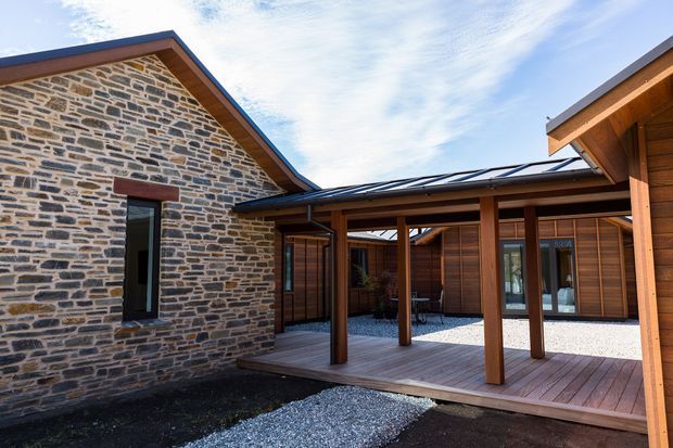 Exterior courtyard view of a low-rise architectural complex combining rough stone masonry walls with warm timber-clad structures. A covered timber post-and-beam walkway and deck frame a gravel courtyard, with pitched roof forms, dark metal roofing, and large glazed doors visible. The image has a natural, refined contemporary-rustic character.
