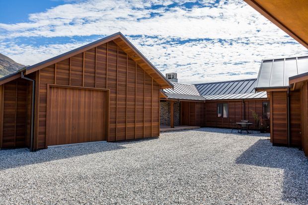 A gravel courtyard framed by multiple timber-clad buildings with steep gabled roofs and a contrasting dark metal roof section. The architecture has a simple lodge or cabin-like composition, with timber garage or entry doors and a small outdoor table. Mountain terrain is visible at the edge of the scene under a bright, cloud-dappled sky.