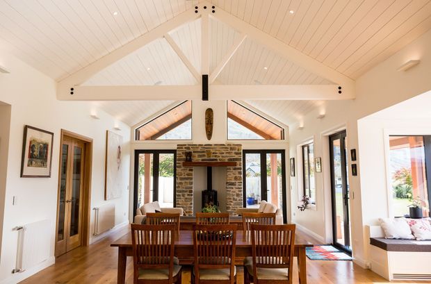 Bright open-plan dining and living space with a large solid timber dining table and chairs centred in front of a rustic exposed brick fireplace. The room features white-painted timber wall and ceiling panelling, prominent exposed white structural beams and rafters, high pitched vaulted ceiling, extensive glazed doors and windows, timber flooring, and a built-in window seat at right.