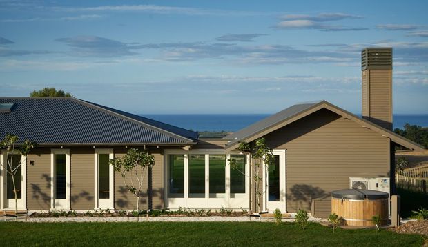 Single-storey coastal home viewed from the rear, with a low-pitched dark metal roof, light weatherboard cladding, extensive glazed doors and windows, a prominent chimney, and a timber outdoor spa or hot tub. The ocean is visible beyond the property.