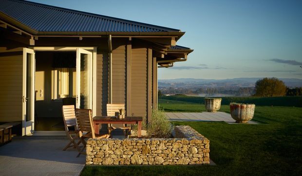 Single-storey rural dwelling or pavilion viewed from a covered outdoor terrace, featuring dark corrugated metal pitched roofing, warm timber weatherboard cladding, exposed eaves, and large glazed hinged doors opening onto the terrace. A timber outdoor table and chairs sit beside a low rough-stone retaining planter wall, with open landscaped grounds and a distant rural landscape beyond.