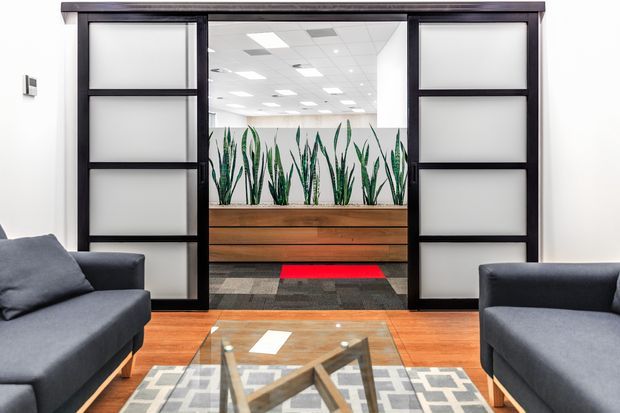 A contemporary office reception or waiting area viewed from a seating zone. Dark charcoal sofas frame a glass coffee table and geometric rug on a warm timber floor. Large black-framed sliding doors with frosted glass panels open toward a bright commercial interior, with a built-in timber planter containing tall architectural plants. The space is clean, bright, and professionally fitted out.