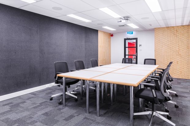 A modern commercial meeting room with a large rectangular conference table surrounded by black office chairs. The room has grey carpet tile flooring, a dark acoustic feature wall, light timber slatted wall panels, white acoustic ceiling tiles with recessed fluorescent lighting, and a glazed entry door with an exit sign.