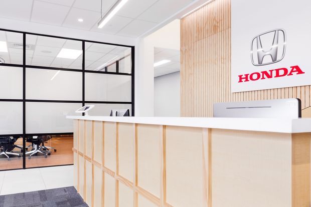 Bright contemporary commercial reception area, likely a Honda dealership or office, with a white reception counter featuring light timber decorative framing, a vertical light-wood slatted feature wall, black-framed glazed office partitions, white acoustic ceiling panels, linear LED lighting, and a prominent Honda logo sign. Office chairs and workspaces are visible behind the glazed partition.