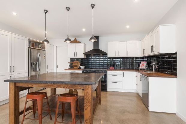 Bright, spacious contemporary kitchen with extensive white shaker-style cabinetry, a large timber-topped island with integrated seating, dark stone-look benchtops, glossy black subway-tile splashback, stainless-steel appliances, and three pendant lights. The room has a warm industrial-meets-contemporary character with strong black, white, timber, and terracotta accents.