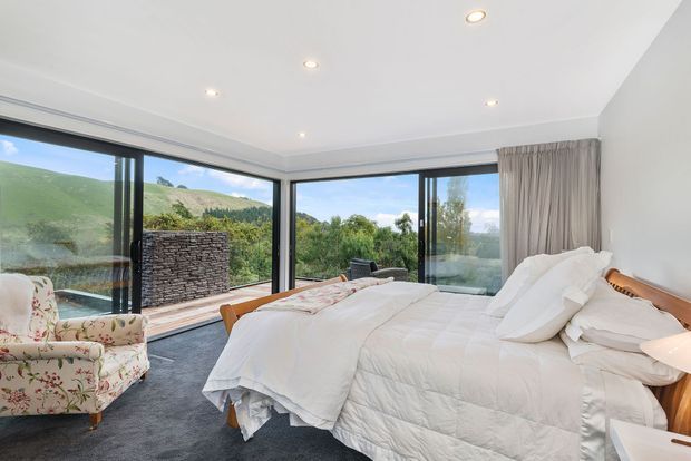 A bright, spacious contemporary bedroom with a large bed dressed in white linens, a floral upholstered armchair, grey carpet, and full-height corner glazing overlooking a green landscape. A dark stacked-stone fireplace or feature wall is visible outside through the glazing, while neutral curtains and a warm timber bed frame add softness and contrast.