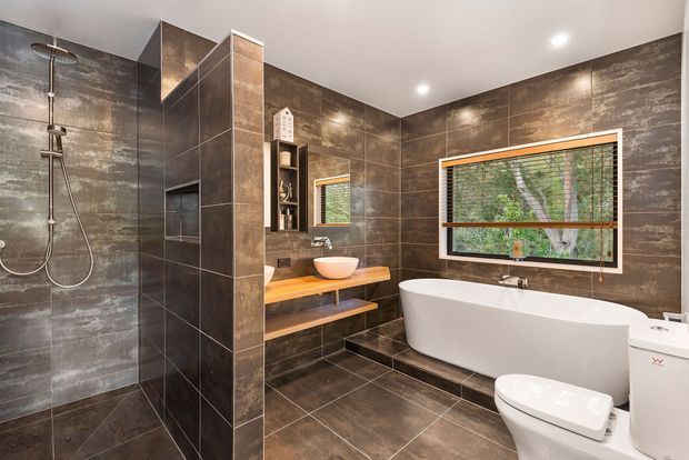 Contemporary bathroom with dark large-format stone-look wall and floor tiles, a frameless glass shower, freestanding white bathtub, wall-mounted timber vanity with vessel basin, toilet, recessed shelving, and a timber-framed window with blinds. Recessed ceiling lighting creates a polished, warm-toned interior.