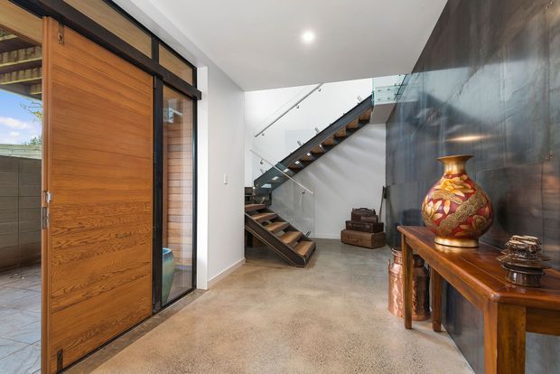 Contemporary entry foyer with a large warm timber pivot door, black metal-framed glazing, polished concrete flooring, and a floating timber stair with glass balustrades. A dark reflective feature wall and timber console with decorative objects add contrast and warmth.