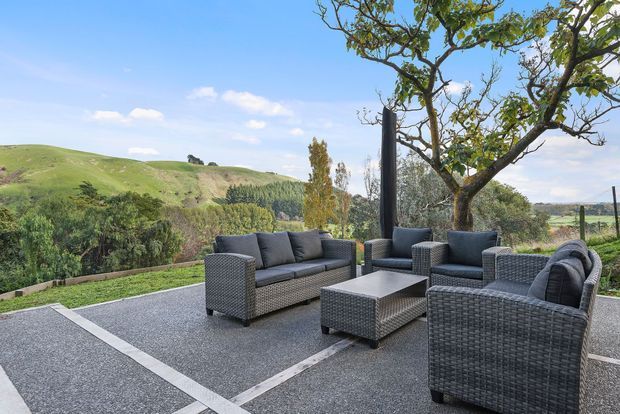 Outdoor lounge area on a spacious paved terrace, furnished with dark woven outdoor sofas and armchairs around a low rectangular coffee table. The terrace overlooks rolling green hills and mature trees, creating a relaxed rural setting.