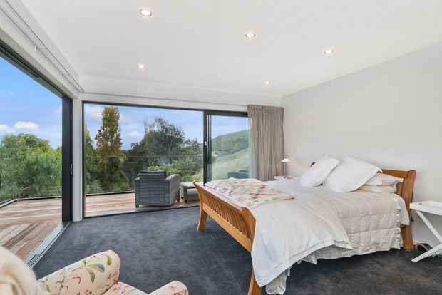 Bright contemporary bedroom with a timber bed, white quilted bedding, dark carpet, and full-height corner glazing opening onto a timber deck and leafy hillside views. A floral upholstered chair and bedside furniture add soft decorative accents, while recessed ceiling lights and neutral curtains complete the room.