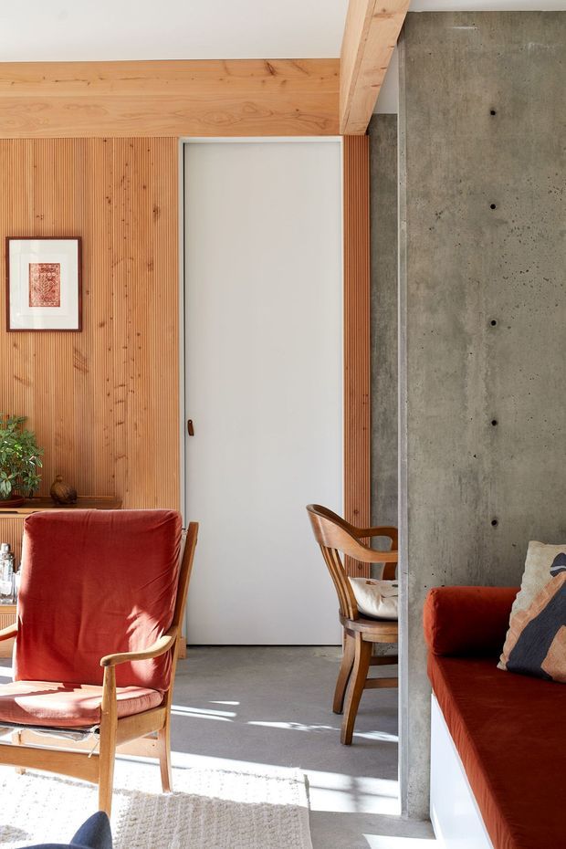 Interior view of a warm, contemporary living space featuring vertical timber wall panelling and exposed timber beams, a smooth concrete structural wall, a white flush door, and mid-century-style timber seating upholstered in deep rust-red fabric. Natural light falls across a neutral rug and concrete floor, with a small framed artwork and potted plant adding detail.