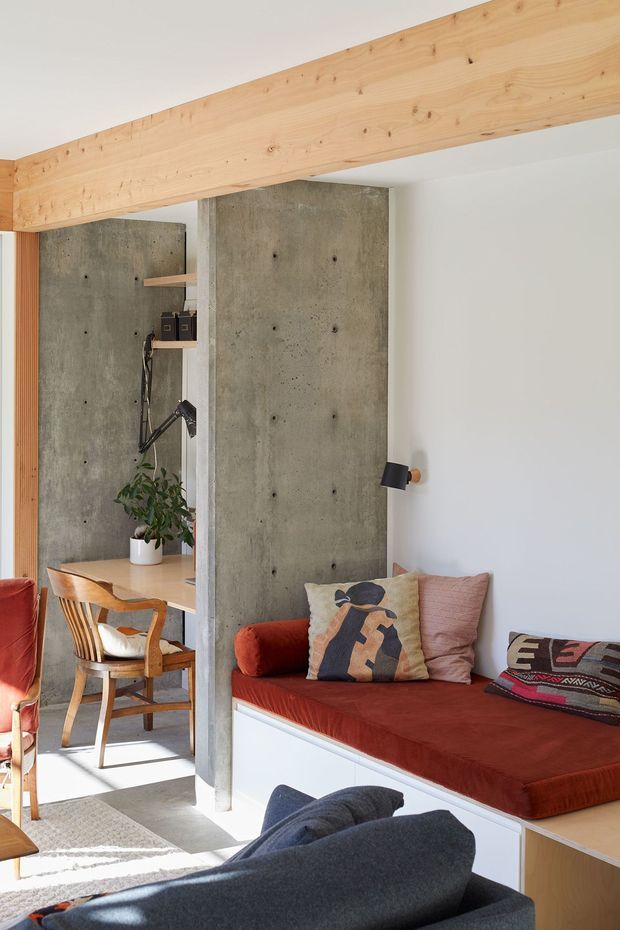 A bright, compact contemporary living room with a built-in window seat or daybed upholstered in rust-red fabric, white storage cabinetry below, and patterned cushions. Exposed concrete feature walls and columns contrast with a large light timber structural beam and timber-framed furniture. A small adjoining desk or study nook is visible to the left, with a black task lamp and potted plant. The room has a warm, curated, mid-century-influenced character with strong natural light.