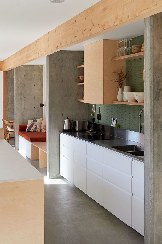 A compact modern kitchen with white flat-panel cabinetry, a dark stone benchtop, integrated sink and cooktop, and open plywood shelving. The kitchen is framed by exposed raw concrete walls and columns, with a large light timber structural beam overhead. A green perforated backsplash adds colour, while a built-in timber seating nook is visible beyond the kitchen.