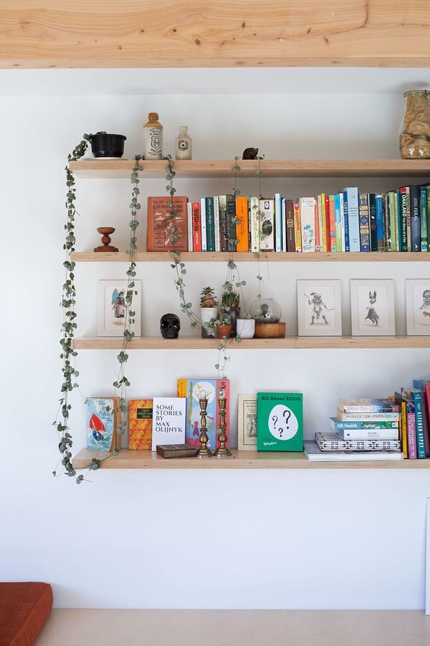 A bright, minimalist built-in wall shelving display with four long light timber shelves mounted against a white wall. The shelves hold colourful books, framed artwork, potted trailing plants, small decorative objects, bottles and ceramics. A timber-clad ceiling or upper wall plane is visible above, with a small portion of a rust-coloured sofa and pale flooring at the bottom of the image.