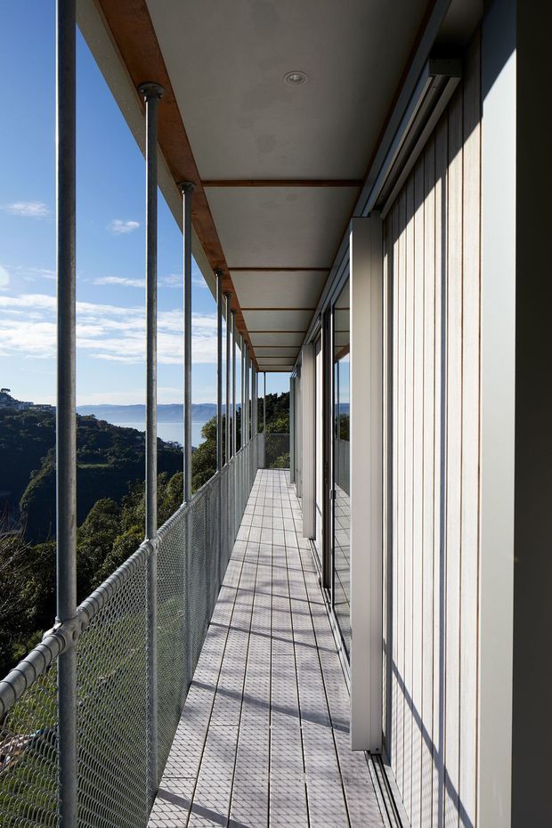 A long, narrow elevated exterior walkway or balcony runs alongside a contemporary building. Slim metal posts support a covered roof canopy, with chain-link safety balustrading along the open edge. The building side has pale vertical cladding, large glazed openings, and dark framing. A wooded, hilly landscape and distant water are visible beyond the railing under clear daylight.
