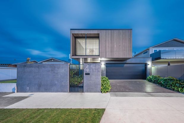 Contemporary two-storey residential facade photographed at dusk, featuring a cantilevered upper volume clad in vertical timber boards, a large framed window with blinds, concrete walls and pillars, a dark garage door, and a gated driveway. Adjacent suburban homes and simple landscaping are visible around the property.