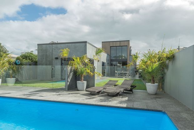 Contemporary single-storey home viewed from a swimming pool, with a flat-roofed geometric facade, dark vertical cladding, extensive glazing, concrete-paved pool deck, synthetic lawn, tropical potted plants and outdoor loungers. The image is taken from the outdoor pool area and shows the rear facade and courtyard-like setting.