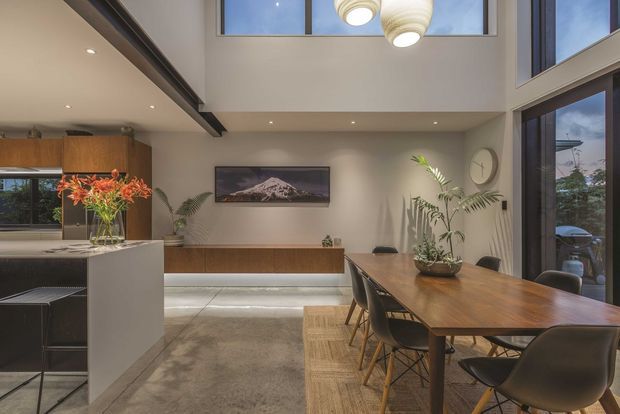 Open-plan contemporary kitchen and dining area with a warm timber dining table, sculptural pendant lights, large windows, a kitchen island with bar seating, timber cabinetry, indoor plants and a double-height ceiling with clerestory glazing.
