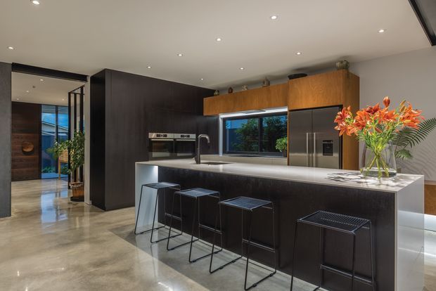 A spacious contemporary kitchen with a large dark island, waterfall-style light stone benchtop, integrated appliances, timber cabinetry, dark full-height joinery, stainless-steel refrigerator, and four minimalist bar stools. Polished concrete or stone-look flooring, recessed ceiling lights, and a glazed opening to the exterior create a refined open-plan setting. A hallway or entry area is visible to the left.