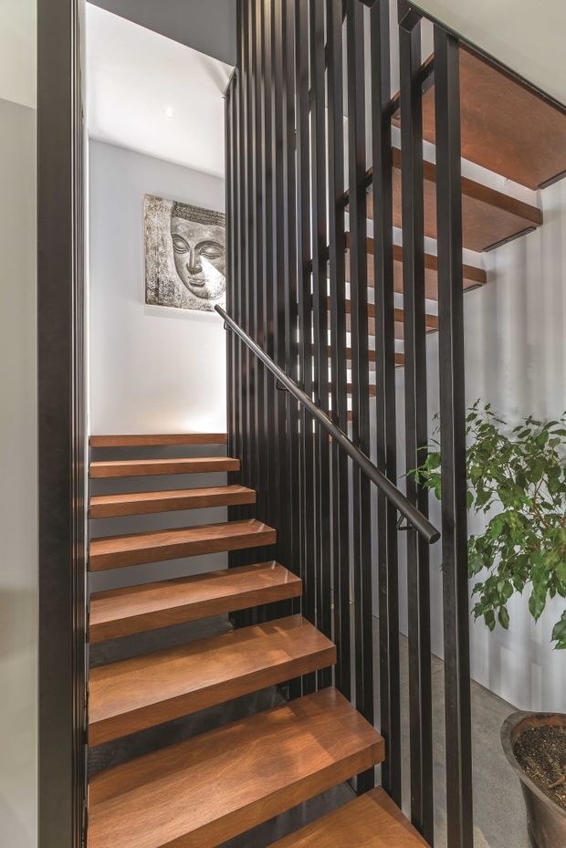 Interior view of a contemporary stairway with warm timber floating treads, dark vertical metal balustrade screening, and a matching dark handrail. White walls, grey flooring, and a potted plant create a clean, refined setting, with artwork visible on the landing wall.