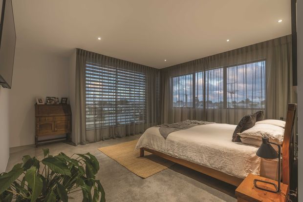 A warm, contemporary bedroom with a low timber bed, neutral bedding, large corner windows fitted with layered curtains and blinds, recessed ceiling lights, a timber chest of drawers, and a bedside table with a lamp. The room has a calm neutral palette and soft natural light, with a large indoor plant in the foreground.