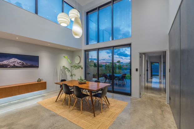 Double-height contemporary dining and living area with a timber dining table, dark upholstered chairs, large woven rug, wall-mounted television, timber media console, indoor plants, and sculptural pendant lights. Full-height glazing opens the room to a covered outdoor setting and distant landscape views, while polished concrete flooring and restrained grey finishes create a clean, refined interior.