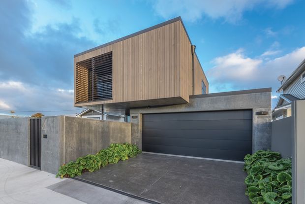 Street-level exterior view of a contemporary multi-level residence with a projecting upper volume clad in vertical timber boards, a deep louvred opening, and a dark integrated double garage. The lower facade uses exposed-looking grey concrete, with a concrete driveway, boundary wall, pedestrian gate, and low planting.