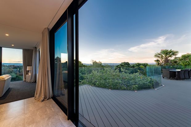 A contemporary indoor-outdoor living space opening onto a spacious grey composite deck. Floor-to-ceiling black-framed glazing and sliding doors connect the interior lounge to the terrace, which features a frameless glass balustrade, outdoor dining furniture, and landscaped greenery beyond. The interior has warm pale flooring, recessed ceiling lights, neutral curtains, and expansive views across the surrounding landscape.