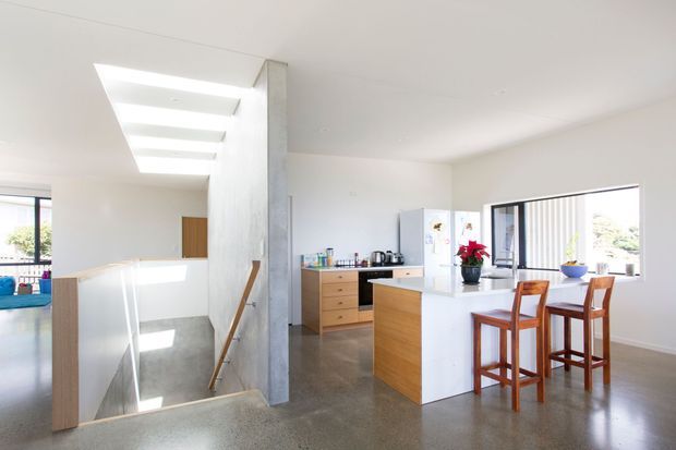 Bright open-plan kitchen and dining area viewed from an upper-level landing. The space features white walls and ceilings, polished grey concrete flooring, pale timber cabinetry, a white kitchen island, and a high clerestory-like skylight over the stairwell. A dining counter with timber bar stools sits beside large windows, while the stair opening and exposed concrete structural column add architectural definition.