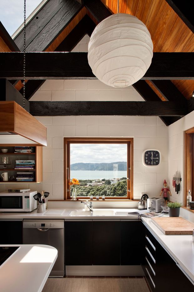 A compact, warm kitchen with black lower cabinetry, white benchtops and white painted block walls. The room features exposed dark timber beams and a timber-lined pitched ceiling, timber-framed windows, open shelving, stainless-steel appliances, and a large white spherical pendant light. A central window provides a prominent view toward a body of water and surrounding hills.