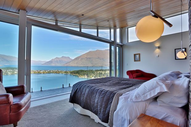 A spacious contemporary bedroom with floor-to-ceiling glazing overlooking a turquoise lake and mountainous landscape. The room features a low-profile bed with grey quilted bedding, pale carpet, warm timber plank ceiling, extensive dark-framed glazing, leather lounge chairs, a red accent chair, and spherical pendant lighting.