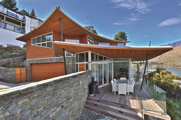 Exterior view of a contemporary multi-level hillside house with dramatic upswept timber roof eaves, warm timber cladding and soffits, extensive glazing, dark stone retaining walls, and a rooftop-style outdoor deck with glass balustrade and dining furniture. A garage is integrated into the lower level, while mountainous terrain and a lake are visible beyond the property.