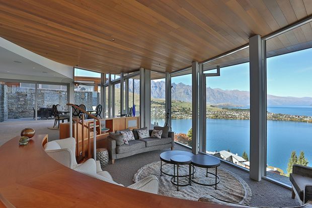 A bright, open-plan living and dining space with expansive floor-to-ceiling glazing overlooking a blue lake and mountainous landscape. The room features a warm timber-lined ceiling, exposed structural columns, a large timber dining table with white chairs, a grey sofa, round coffee tables, and a textured circular rug. The glazing wraps around the room to maximise the panoramic view.