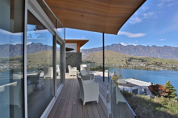 A modern elevated terrace overlooking a blue lake and rugged mountain landscape. The outdoor deck features timber flooring, a deep timber-lined soffit, frameless glass balustrades, full-height glazing, minimalist outdoor lounge chairs, and a stone-clad privacy wall. The view and architecture create a refined, airy connection between the interior and landscape.