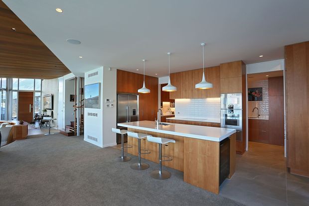 Open-plan contemporary kitchen and living area with extensive warm timber cabinetry, a large white island with integrated sink and breakfast-bar seating, stainless-steel appliances, pale tiled splashback, and multiple pendant lights. The kitchen opens to a carpeted living zone with tall glazing and a timber-lined ceiling feature.