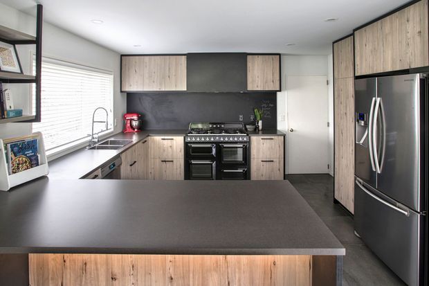 A contemporary kitchen viewed from behind a large island, featuring wood-grain cabinetry, dark charcoal benchtops and splashback, a freestanding stainless-steel range cooker, integrated shelving, a sink beneath a wide window, and a large stainless-steel refrigerator. The space has a clean, functional layout with recessed ceiling lights and a neutral palette.