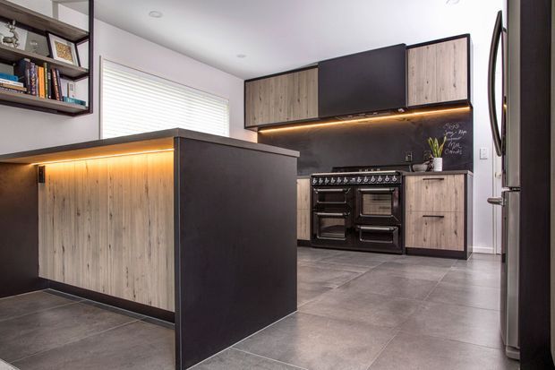 A contemporary kitchen featuring a dark central island with warm under-cabinet lighting, light timber-look cabinetry, matte dark upper cabinets, a large freestanding range cooker, charcoal splashback with handwritten notes, grey tiled flooring, and stainless-steel refrigeration. Open shelving with books and framed objects is visible at the left.