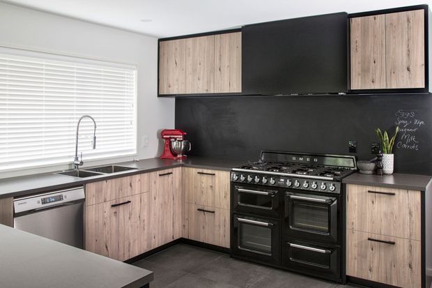 A contemporary L-shaped kitchen with light wood-grain cabinetry, matte black upper cabinets and splashback, dark benchtops, and a large black freestanding dual-oven range. The space includes a stainless-steel dishwasher and sink beneath a wide window with white blinds, plus a red stand mixer and chalkboard-style writing on the black splashback.