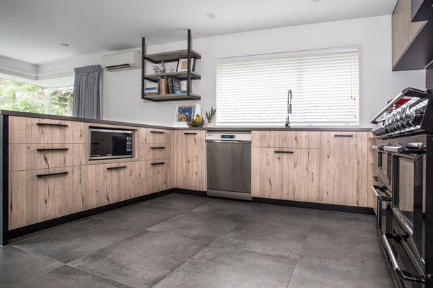 A contemporary kitchen with an L-shaped run of light timber-grain cabinetry, black handles and dark benchtops. The room includes a stainless-steel dishwasher, integrated microwave, large freestanding range cooker, undermount sink, open black-framed shelving, white walls, window blinds and large-format grey floor tiles.