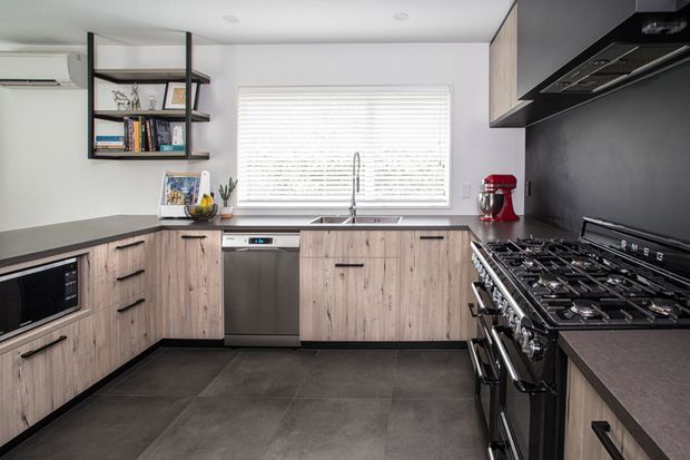 Modern U-shaped kitchen with light wood-grain cabinetry, dark stone-look benchtops, a black feature wall and splashback, stainless-steel dishwasher, large freestanding gas range, under-cabinet microwave, and open shelving. A wide window with white blinds provides natural light.