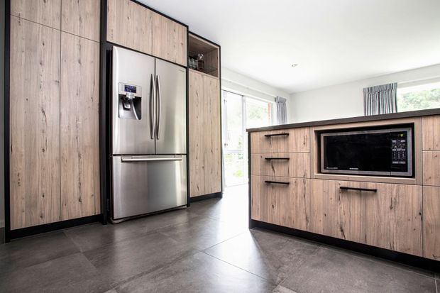 A contemporary kitchen with full-height light timber-look cabinetry, black edging and handles, stainless-steel French-door refrigerator, integrated microwave, and dark grey tiled flooring. A bright windowed area is visible beyond the cabinetry.