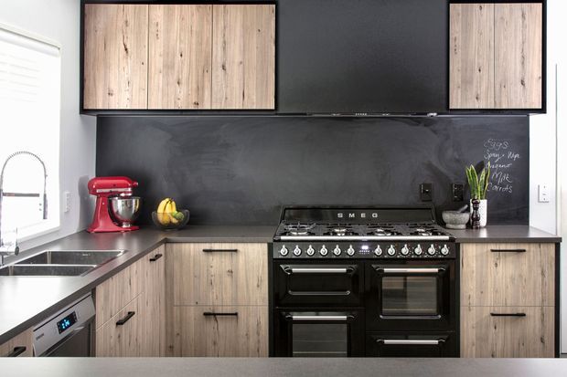 A compact contemporary kitchen with a U-shaped layout, light wood-grain cabinetry, a dark charcoal backsplash and matching dark benchtops. A large black freestanding range cooker is centered beneath upper cabinets, with a red stand mixer, sink, dishwasher, fruit bowl, plant, and chalkboard-style notes adding functional and decorative details.
