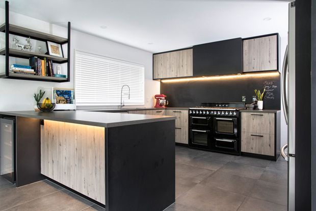 Contemporary kitchen with a large central island, dark cabinetry, light timber-look cabinet fronts, integrated under-cabinet lighting, and a prominent black range cooker. Open wall shelving, white window blinds, and large-format grey floor tiles complete the modern, high-contrast scheme.